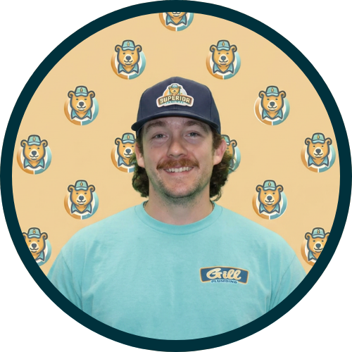 Smiling man in a teal Gill Plumbing shirt and matching cap, posing in front of a bear-patterned backdrop with circular logo repeats.