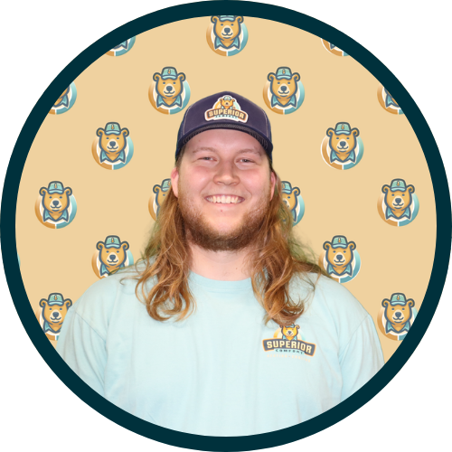 Portrait of a smiling man with long hair wearing a light blue shirt and a cap, standing against a beige background with a repeating bear logo pattern.