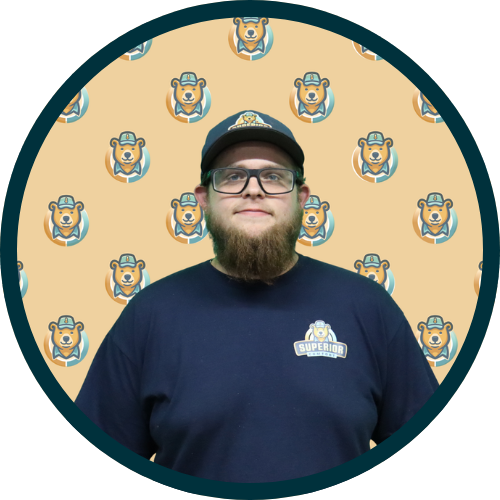 Portrait of a man with glasses and a full beard wearing a navy shirt and cap, standing before a beige backdrop with a repeating dog logo pattern.] ,first person