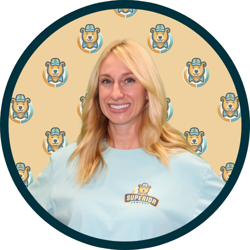 Portrait of a smiling woman with shoulder-length blonde hair, wearing a light blue shirt with a bear/logo emblem, standing against a beige background patterned with bear mascots.